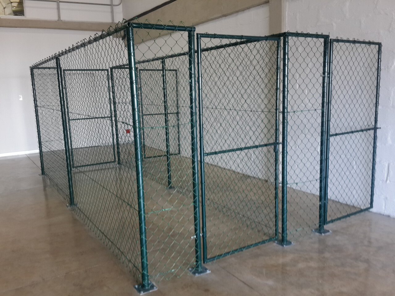 Green plastic-coated chain-link partitioning inside a warehouse, floor-mounted on base plates, forming storage cages.