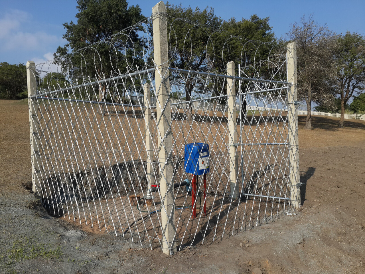 A small enclosure of galvanised diamond-mesh fencing on concrete posts, topped with razor coil, protecting a blue pump installation on a rural site near Darnall.
