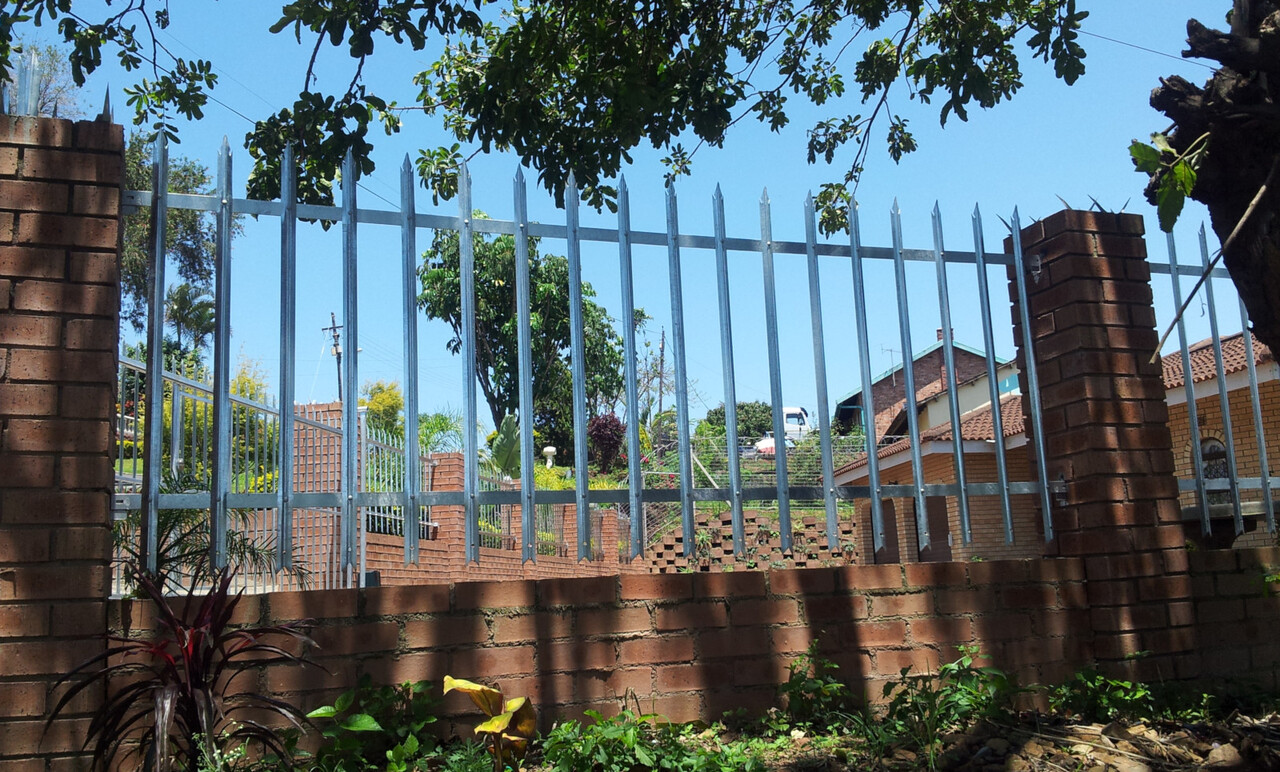 Galvanised steel palisade fencing with pointed pales, mounted on top of a red-brick boundary wall with brick pillars on a Verulam suburban street.