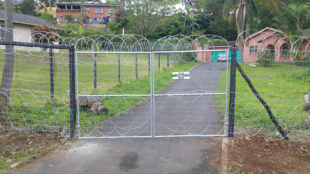 A galvanised weld-mesh double swing gate topped with razor coil on a residential driveway, Durban hillside houses behind — a completed Verulam Fencing job.